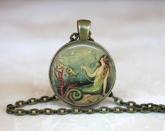 Op de afbeelding: Een ronde hangerketting met een bronskleurige ketting. De hanger toont een vintage illustratie van een zeemeermin in een onderwaterwereld met vissen en koraal. De zeemeermin houdt een parelketting vast.