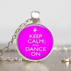 May include: A silver-toned necklace featuring a round pendant with a bright pink background. The pendant has the words "KEEP CALM and DANCE ON" in white, along with a ballerina silhouette. The necklace is a dance-themed accessory.