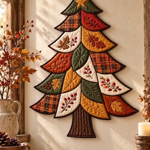 Op de afbeelding: Een gewatteerde wandhanger in de vorm van een kerstboom, opgebouwd uit stoffen segmenten met herfstpatronen, waaronder ruiten, effen kleuren en geborduurde blad- en bessenmotieven, bekroond met een kleine stoffen ster.