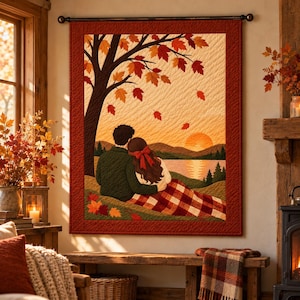 Op de afbeelding: Een rechthoekige wandquilt met een scène van twee personen die in de herfst onder een boom zitten. Het ontwerp bevat een zonsondergang boven bergen, een meer, vallende oranje en rode bladeren en een rood-witte geruite deken.