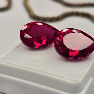 Puede incluir: Dos rubíes facetados en forma de pera, de color rojo intenso y gran claridad. Las piedras preciosas se muestran una al lado de la otra sobre una almohadilla de presentación blanca y cuadrada con una superficie suave y texturizada.