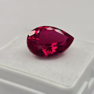 Può includere: Una pietra preziosa sfaccettata a forma di pera con un colore rosso intenso e vibrante. La pietra presenta un taglio brillante con molteplici superfici riflettenti che catturano la luce, mostrando una qualità chiara e trasparente.