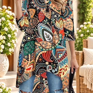 May include: A tunic top featuring a black base with a vibrant, multi-colored paisley and leaf pattern, three-quarter length sleeves, a round neckline with a small button placket, and a high-low hemline paired with distressed blue denim jeans.