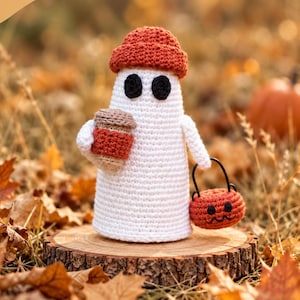 May include: A white crochet ghost figure wearing a rust-colored beanie, holding a tan and rust-colored coffee cup in one hand and a small orange jack-o'-lantern bucket in the other. The ghost has two black circular eyes.