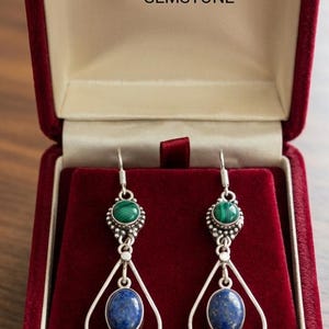 May include: A pair of drop earrings featuring a round green malachite stone set above an oval blue lapis lazuli stone, encased in silver metal frames. The text LAPIS AND MALACHITE GEMSTONE is printed above the earrings in the box.