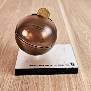 Puede incluir: Una hucha esférica de color bronce con acabado metálico, que tiene una moneda insertada en la ranura superior. Está montada sobre una base rectangular blanca con el texto negro "JOURNEE MONDIALE DE L'EPARGNE 1978" impreso en la parte frontal.