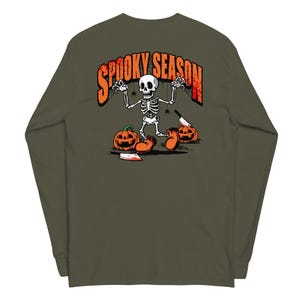 Peut inclure: Un t-shirt à manches longues vert olive avec un motif au dos. Le dessin représente un squelette de dessin animé portant de grandes chaussures orange, entouré de deux citrouilles, deux couperets et des araignées, avec le texte SPOOKY SEASON.