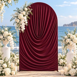 Op de afbeelding: Een hoge, boogvormige achtergrondstandaard bedekt met diepbordeauxrode stof met verticale, geplooide draperieën. De boog is versierd met een groot arrangement van witte rozen, orchideeën en groen aan de linkerbovenkant.