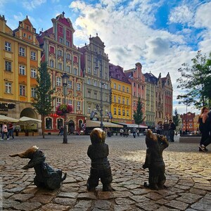Może przedstawiać: Okładka przewodnika turystycznego z tytułem BRESLAVIA IN 48 ORE, La Guida Definitiva tra Gnomi, Isole e Lanterne a Gas, Edizione Low Budget & Smart, przedstawiająca trzy brązowe figurki krasnali na brukowanym placu przed kolorowymi kamienicami.