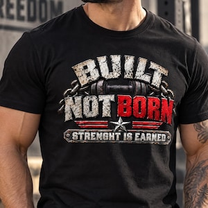 Könnte beinhalten: Ein schwarzes Kurzarm-T-Shirt mit einem Grafikdruck, der den Text "BUILT NOT BORN" in abgenutzter weißer und roter Schrift, ein Hantelsymbol, einen silbernen Stern und den Text "STRENGHT IS EARNED" auf einem metallartigen Banner zeigt.