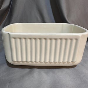 Op de afbeelding: Een rechthoekige, gebroken witte keramische plantenbak of opbergbak met een gladde binnenkant en een decoratieve buitenkant met verticale ribbels aan de zijkanten. De bak heeft afgeronde hoeken en een egale, glanzende afwerking.