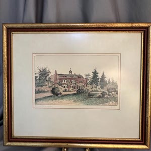 May include: A framed print featuring a detailed etching of a multi-story brick house with a cupola, surrounded by trees and shrubs. The artwork is set within a cream-colored mat and a decorative gold and dark red wooden frame with a textured finish.