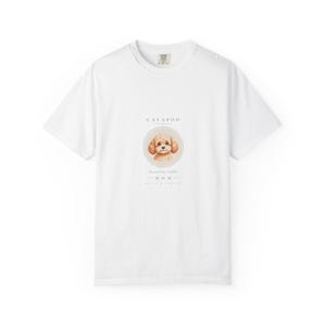 May include: A white short-sleeve t-shirt featuring a central graphic of a light brown Cavapoo dog inside a circular frame. The text above the dog reads CAVAPOO, and the text below reads Powered by Cuddles M O M with additional small text.