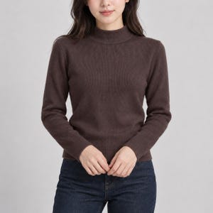 Könnte beinhalten: Ein dunkelbrauner Langarmpullover mit gerippter Strickstruktur und Stehkragen. Das Kleidungsstück wird mit einer dunkelblauen Jeans kombiniert, was ein schlichtes und funktionelles Outfit mit langen Ärmeln und einer körpernahen Passform ergibt.