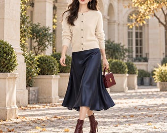 May include: A beige long-sleeved button-down cardigan paired with a navy blue satin midi skirt, a small dark red handbag, and dark red heeled ankle boots. The outfit features a soft-textured knit top and a smooth, flowing skirt.