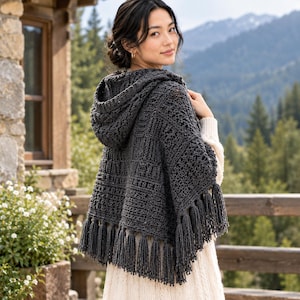 May include: A dark charcoal gray hooded crochet poncho featuring a textured geometric pattern and long fringe trim along the bottom edge. The garment is draped over the shoulders and includes a hood made of the same dark gray yarn.