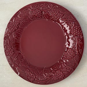 May include: A round, deep burgundy ceramic plate featuring a raised, embossed border design of grape clusters and vine leaves. The center of the plate is smooth and plain, while the rim is decorated with a continuous pattern of fruit.