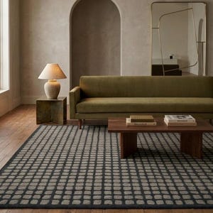 Könnte beinhalten: Ein rechteckiger Teppich mit einem dunkelgrauen und hellgrauen Gittermuster. Der Teppich liegt in einem Raum mit einem langen olivgrünen Sofa, einem hölzernen Couchtisch mit zwei Büchern, einem Beistelltisch mit Lampe und einem hohen Spiegel.