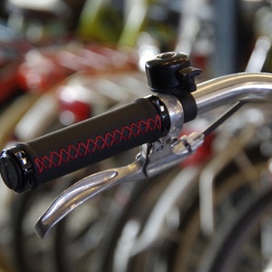 May include: Close-up of a black leather bicycle handlebar grip with red stitching. The grip is attached to a silver handlebar with a silver brake lever.