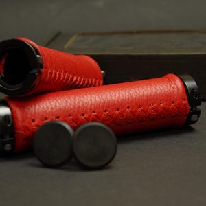 May include: Two red leather bicycle handlebar grips with black end caps and stitching. The grips are on a black surface.