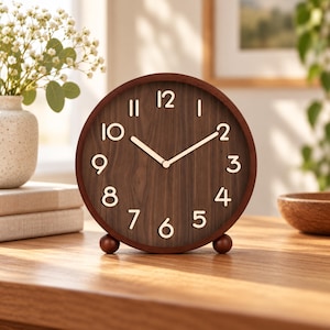 May include: A round analog table clock with a dark brown wood grain face and a matching circular frame. The clock features raised, light-colored numerals from 1 to 12 and two matching hands. It stands on two small, spherical brown feet.
