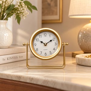 May include: A gold-toned metal table clock with a circular frame and a white face featuring black Arabic numerals and hands. The clock is mounted on a U-shaped stand that allows for rotation, with the word QUARTZ printed on the dial.