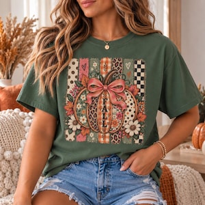 May include: An olive green t-shirt featuring a central graphic of a pumpkin adorned with a pink bow and the word FALL. The design includes vertical stripes with polka dots, leopard print, plaid, and floral patterns, surrounded by autumn leaves.