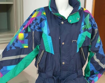 May include: A navy blue Fila ski suit featuring a high collar, long sleeves, and a belted waist. The garment is decorated with geometric patterns in shades of bright green, light blue, purple, and yellow, with silver-toned metal zippers.