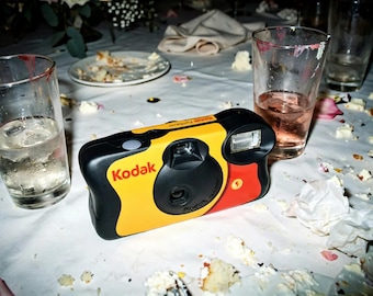 May include: A Kodak FunSaver disposable camera featuring a black, yellow, and red plastic body with a built-in flash, a central lens, and a shutter button on top. The brand name Kodak is printed in red on the front of the camera body.
