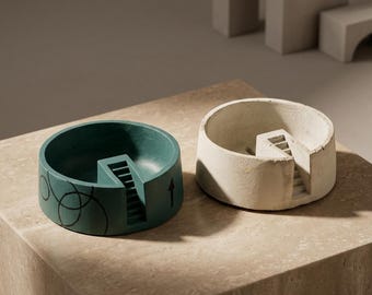 May include: Two circular concrete bowls, one dark teal and one off-white, each featuring a small internal staircase structure. The teal bowl displays black line drawings of overlapping circles and a vertical arrow on its exterior side.