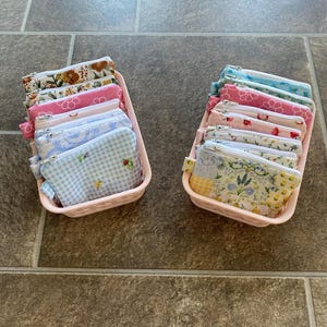 May include: Two light pink plastic baskets filled with small zippered pouches. The pouches feature various patterns including blue gingham, floral prints, and pink daisy designs. Each pouch has a silver metal zipper closure along the top edge.