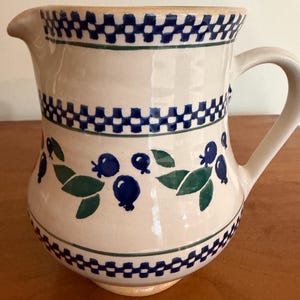 May include: A cream-colored ceramic pitcher featuring a hand-painted blue and green botanical pattern of berries and leaves, accented by horizontal bands of a blue and white checkered design around the rim and the lower body.
