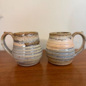 May include: Two handmade ceramic mugs with a rounded, bulbous shape and a sturdy handle. The mugs feature a textured, ribbed surface with a glaze that transitions from a warm, sandy beige at the base to a darker, mottled brown at the rim.