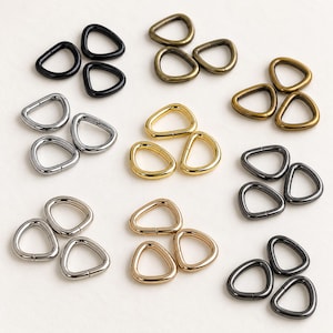 May include: A collection of metal triangle rings arranged in small groups, featuring various metallic finishes including matte black, antique brass, polished gold, silver, gunmetal, and light gold, each with a smooth, rounded triangular shape.