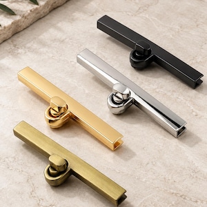 May include: Four rectangular metal bag locks arranged diagonally. Each lock features a central twist-turn mechanism and a long, slender bar. The set includes finishes in matte black, polished silver, shiny gold, and an antique brass tone.