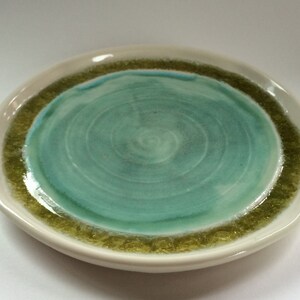 May include: A handmade ceramic plate with a unique design. The plate features a light beige rim, a textured olive green band, and a swirling turquoise center. The plate's design evokes a sense of nature and artistry.