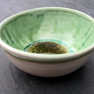 May include: A handmade ceramic bowl with a light green interior and a cream-colored exterior. The bowl's interior features a textured, dark green and blue design at the base. The rim has a darker green trim, adding a touch of contrast to the piece.