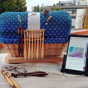 Może przedstawiać: Niebiesko-biała poduszka w tkaninie leży w wiklinowym koszu.  Drewniane szpulki są ułożone w rzędzie przed koszem.  Tablet z ekranem wyświetlającym instrukcje, jak przygotować szpulki, stoi na stole. Tekst na ekranie brzmi "Jak przygotować szpulki".
