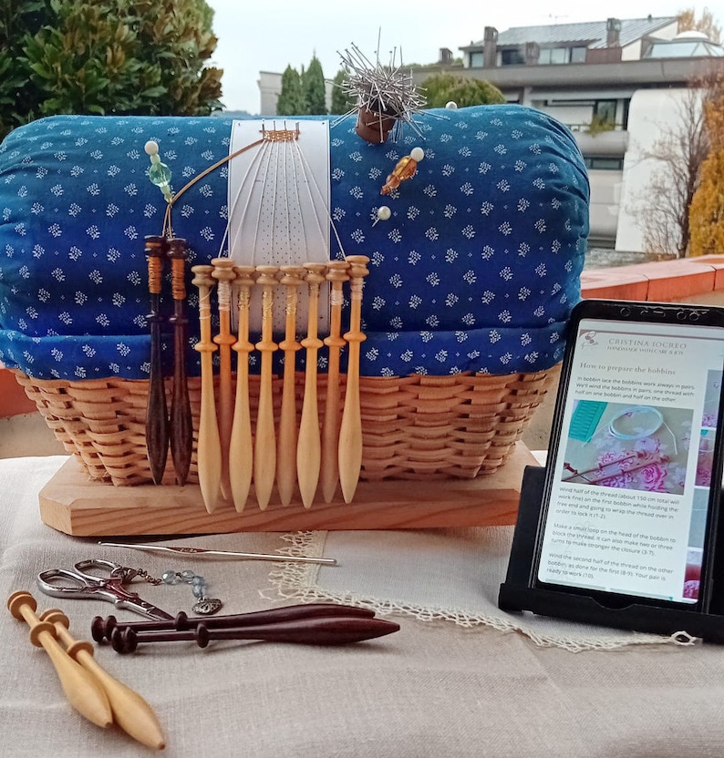 Może przedstawiać: Niebiesko-biała poduszka w tkaninie leży w wiklinowym koszu.  Drewniane szpulki są ułożone w rzędzie przed koszem.  Tablet z ekranem wyświetlającym instrukcje, jak przygotować szpulki, stoi na stole. Tekst na ekranie brzmi "Jak przygotować szpulki".
