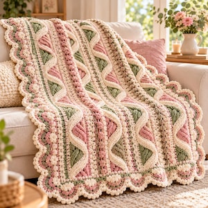 May include: A crocheted blanket featuring a textured pattern of cream, sage green, and dusty rose yarn. The design includes raised cable stitches, bobble stitch rows, and a scalloped edge with small flower-like motifs along the border.