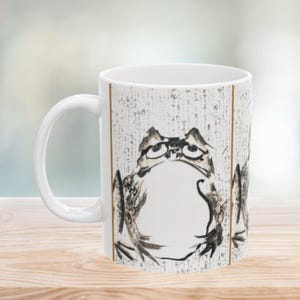 May include: White ceramic mug featuring a detailed ink drawing of a frog with a contemplative expression. The frog is depicted in shades of black and brown against a background of faded text. The mug has a white handle and a vertical brown stripe.