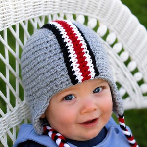 May include: A gray crocheted beanie with ear flaps, featuring a central vertical stripe in black, white, and red. The hat includes braided ties at the bottom of each ear flap, incorporating the same black, white, and red color scheme.