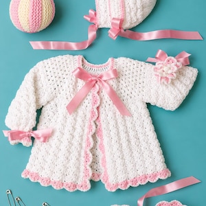 May include: A white crocheted baby set featuring a long-sleeve cardigan with pink ribbon ties, a matching bonnet with pink ribbons, a pair of booties with pink accents, a small flower accessory, a striped ball, and a pincushion with silver safety pins.
