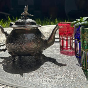 Peut inclure: Une théière en métal argenté avec un motif en relief orné, un bouton en forme d'oiseau sur le couvercle et une anse courbée. À côté se trouvent trois verres rouge, bleu et vert, chacun doté d'une bordure décorative dorée.