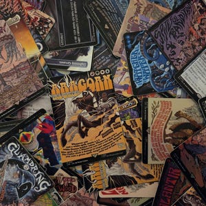 Puede incluir: Una colección de cartas coleccionables rectangulares con diversas ilustraciones de fantasía y texto. Una carta central muestra una figura a caballo con el texto Aragorn, The Uniter. Otras cartas visibles incluyen títulos como Glamdring, The Watcher in the Water y Spiteful Bandit, todas con ilustraciones detalladas y texto relacionado con el juego.