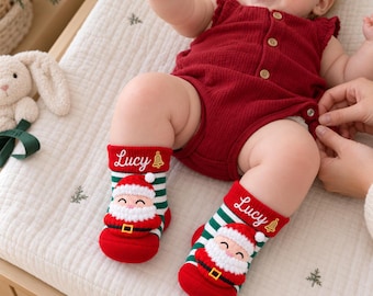 May include: A pair of baby socks featuring green and white horizontal stripes, a red toe, and a red cuff with the name Lucy and a gold bell icon embroidered on it. A 3D plush Santa Claus figure is attached to the front of each sock.
