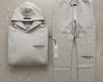 Könnte beinhalten: Ein hellgraues, meliertes Set aus Kapuzenpullover und Jogginghose. Der Kapuzenpullover hat einen schwarzen rechteckigen Aufnäher auf der Kapuze und den Schriftzug ESSENTIALS FEAR OF GOD auf der Brust. Die Jogginghose hat einen Kordelzug.