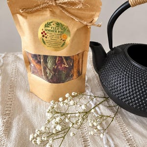 以下が含まれることがあります： Yule Simmer Potと書かれたラベル付きの茶色の紙袋。中には乾燥したオレンジ、クランベリー、シダー、セージ、アニス、クローブが入っており、麻紐で結ばれています。横には黒い鋳鉄製のティーポットと、白いカスミソウの小枝が添えられています。