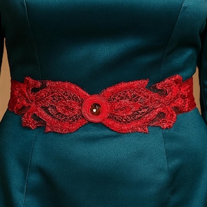 Puede incluir: Una mujer con un vestido color verde azulado con cuello barco y mangas tres cuartos. El vestido se ciñe a la cintura con un cinturón de encaje rojo adornado. El fondo está suavemente iluminado, sugiriendo un ambiente interior.