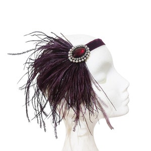 May include: A burgundy velvet headband with a large red rhinestone and pearl embellishment, accented with a long plume of burgundy feathers.